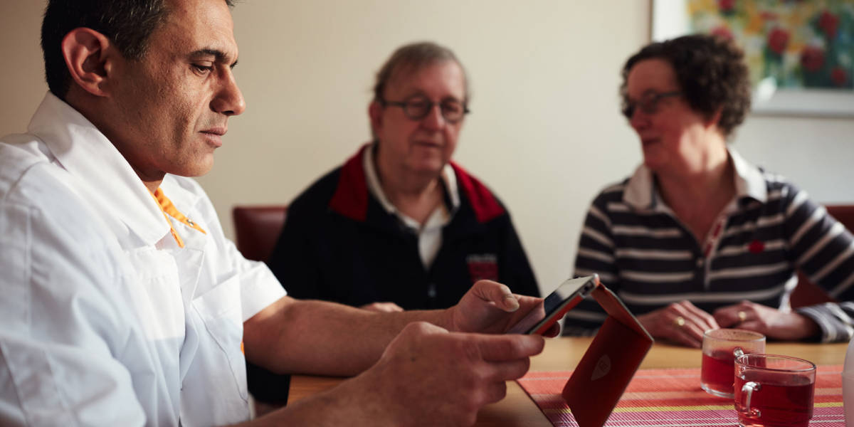 Verpleegkundige bespreekt aan tafel met twee mensen informatie op een tablet